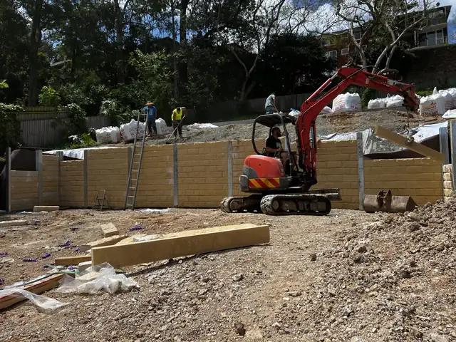 Crew and excavator building a sandstone-look concrete sleeper wall on a sloping site
