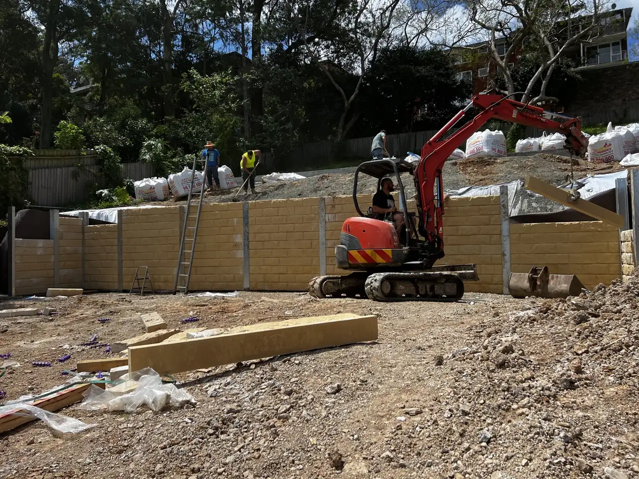 Crew and excavator building a sandstone-look concrete sleeper wall on a sloping site